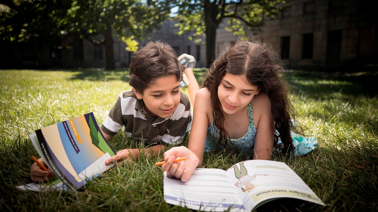 A boy and a girl lying on the grass working on junior ranger books.