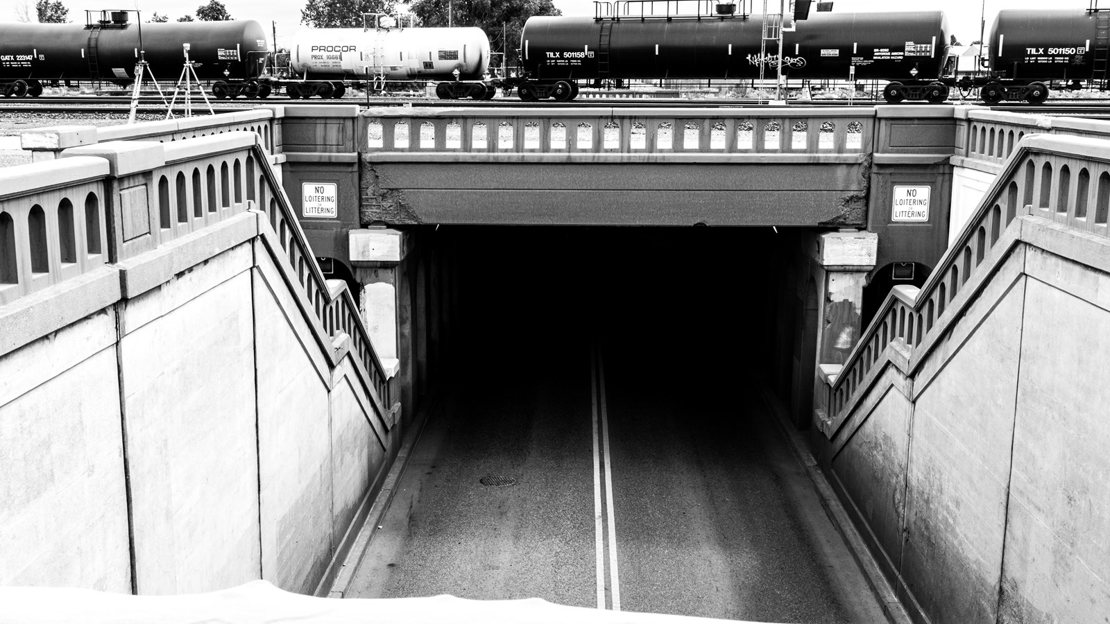 Black and white image of a bridge with multiple stairways. Beneath the bridge is a darkened roadway.