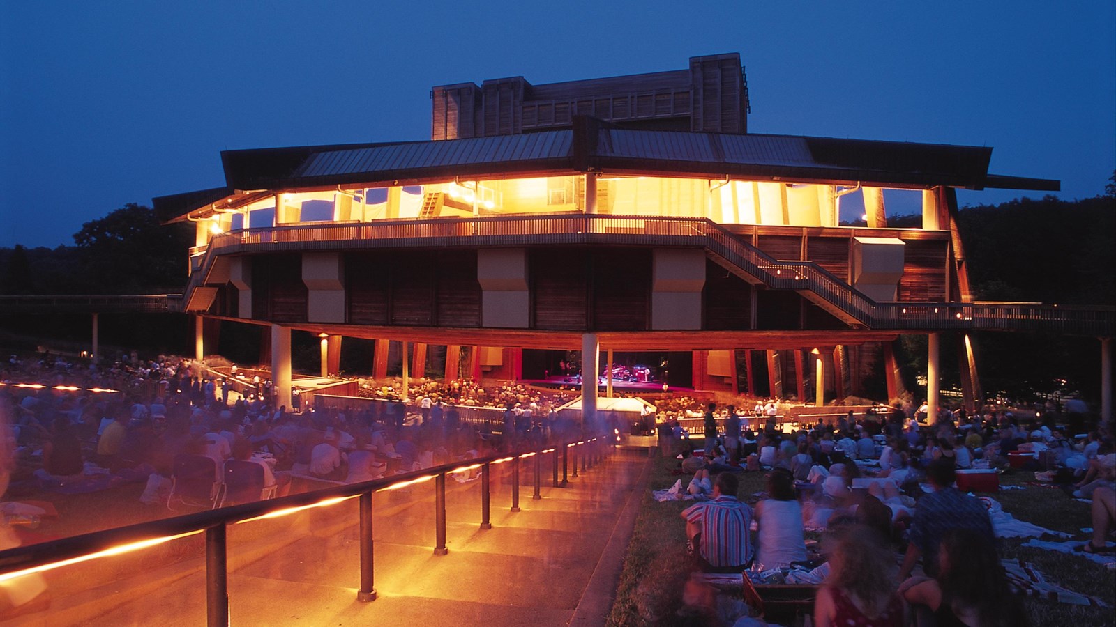 An illuminated wooden stage area with several patrons sitting in a grassy area around it.