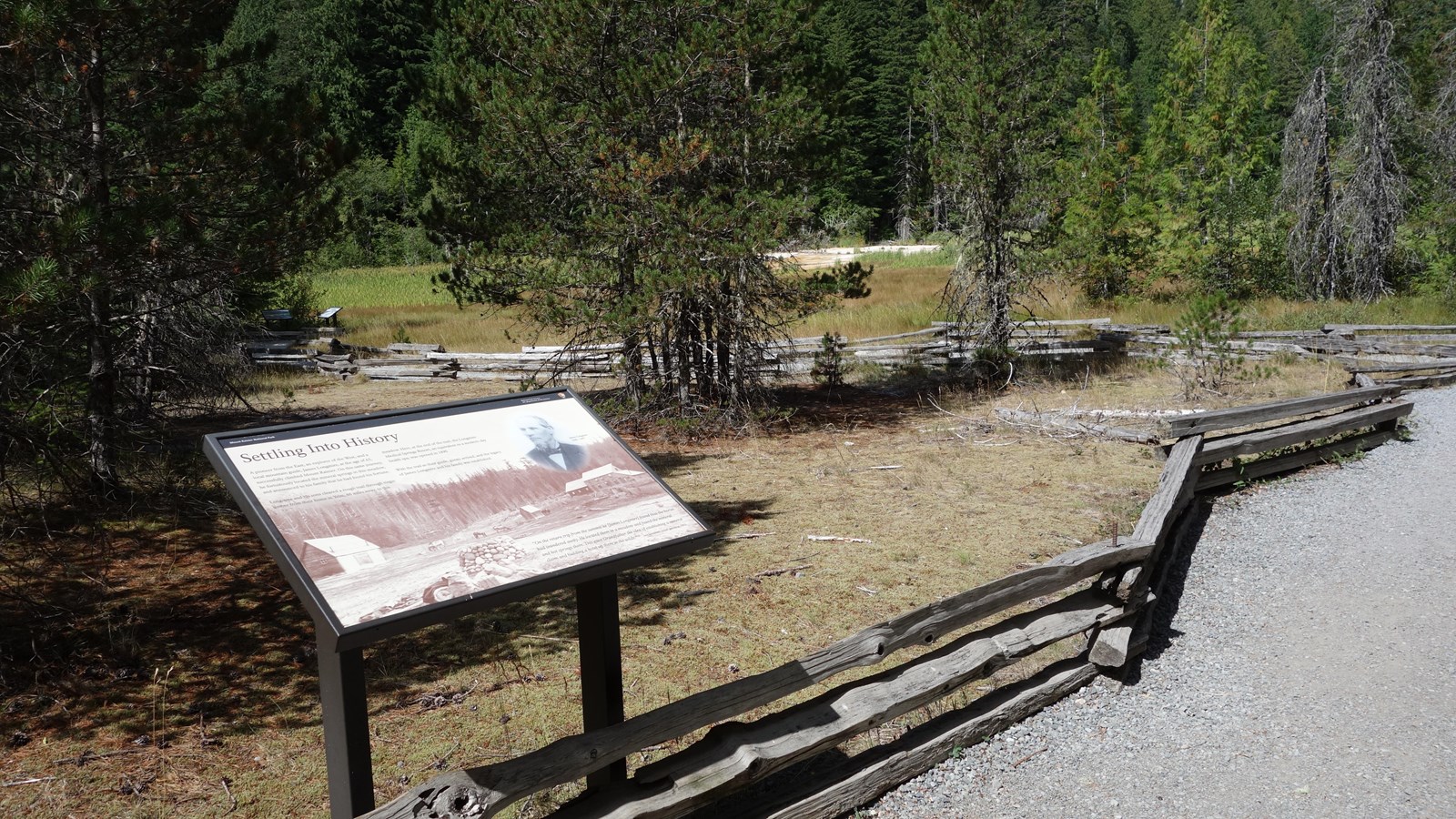 A sign in a metal stand next to trail bordered by a split-rail fence and patches of meadow.