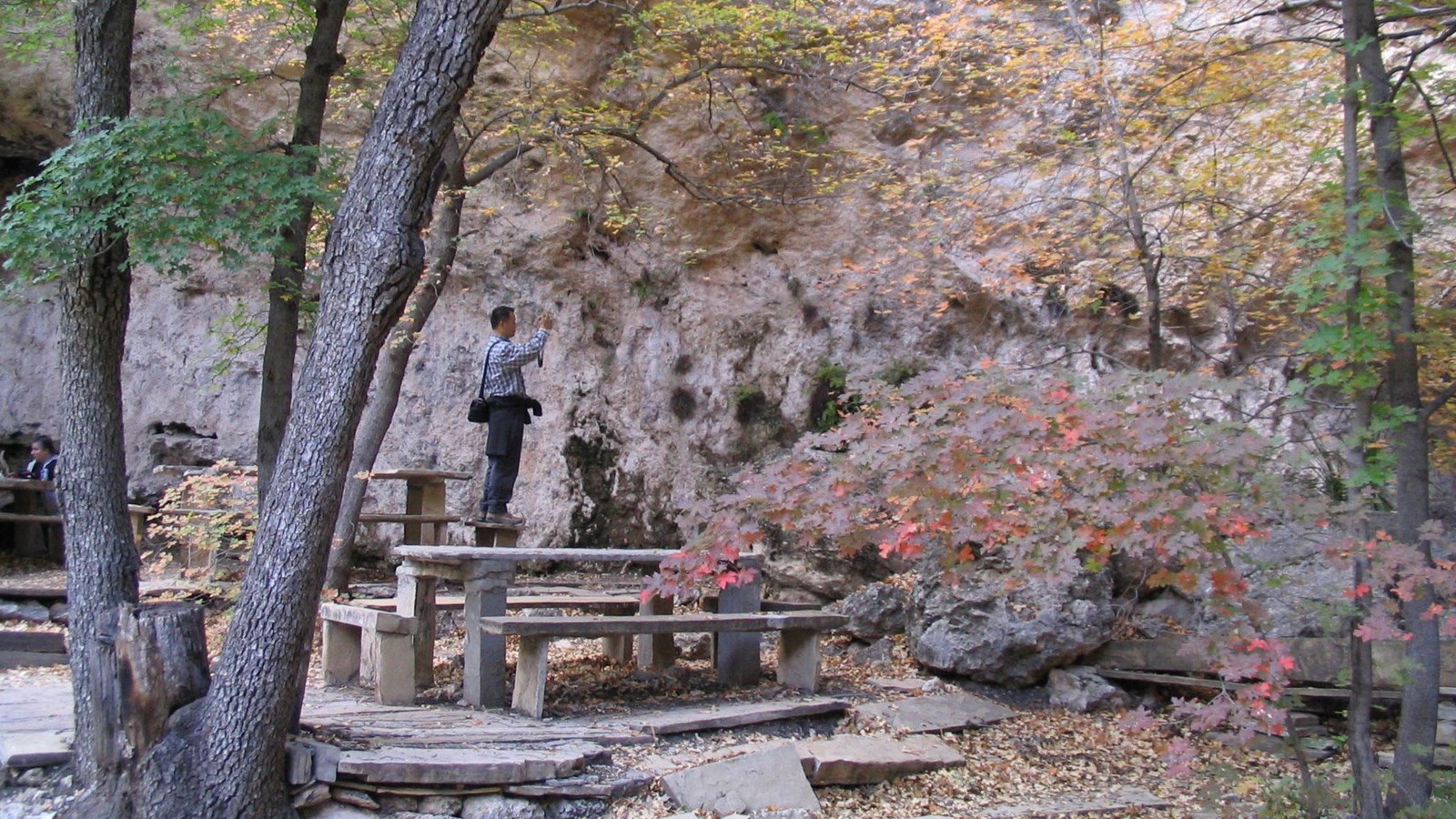 A man use a camera near an open cave feature with stone benches and tables.