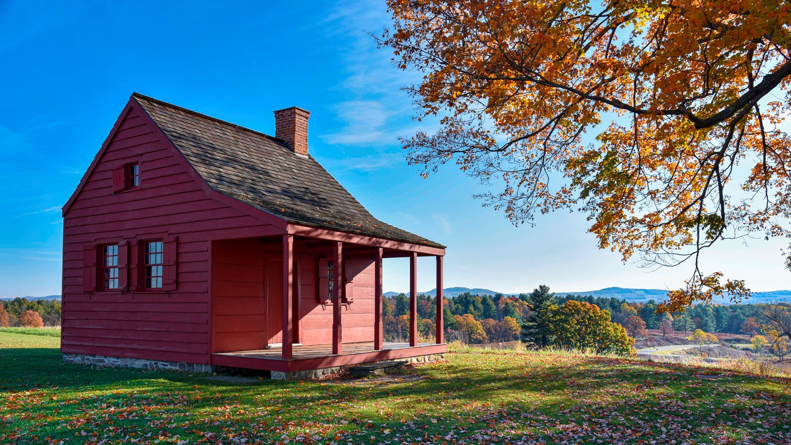 Single story red house with a covered porch, mowed grass in the foreground with fallen leaves