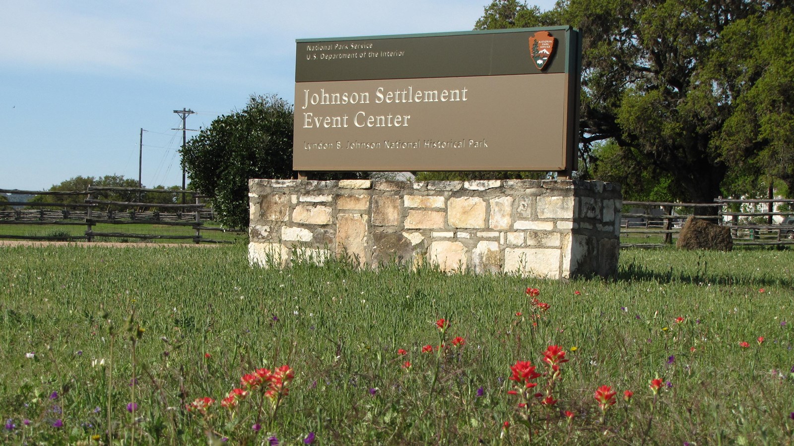 A brown sign sits on a stone base surrounded by green grass and wildflowers.