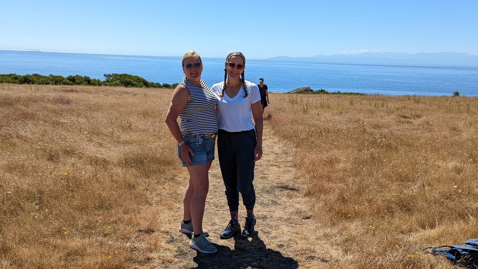 Color photo of 2 hikers on a trail beside grassland. Behind them is the ocean
