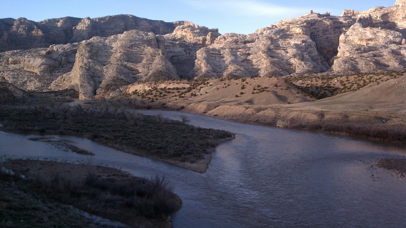 A large river flows in late evening shadow as sun hits a large, rocky mountain in the distance.