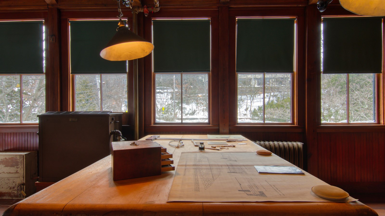 Large wooden table with tools and papers on top, with stools around it. 