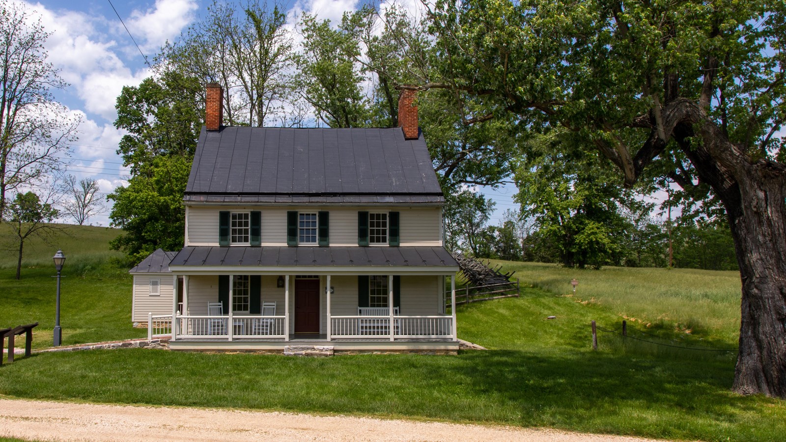 A tan house with green shutters, two chimneys, and a front porch sits among green grass