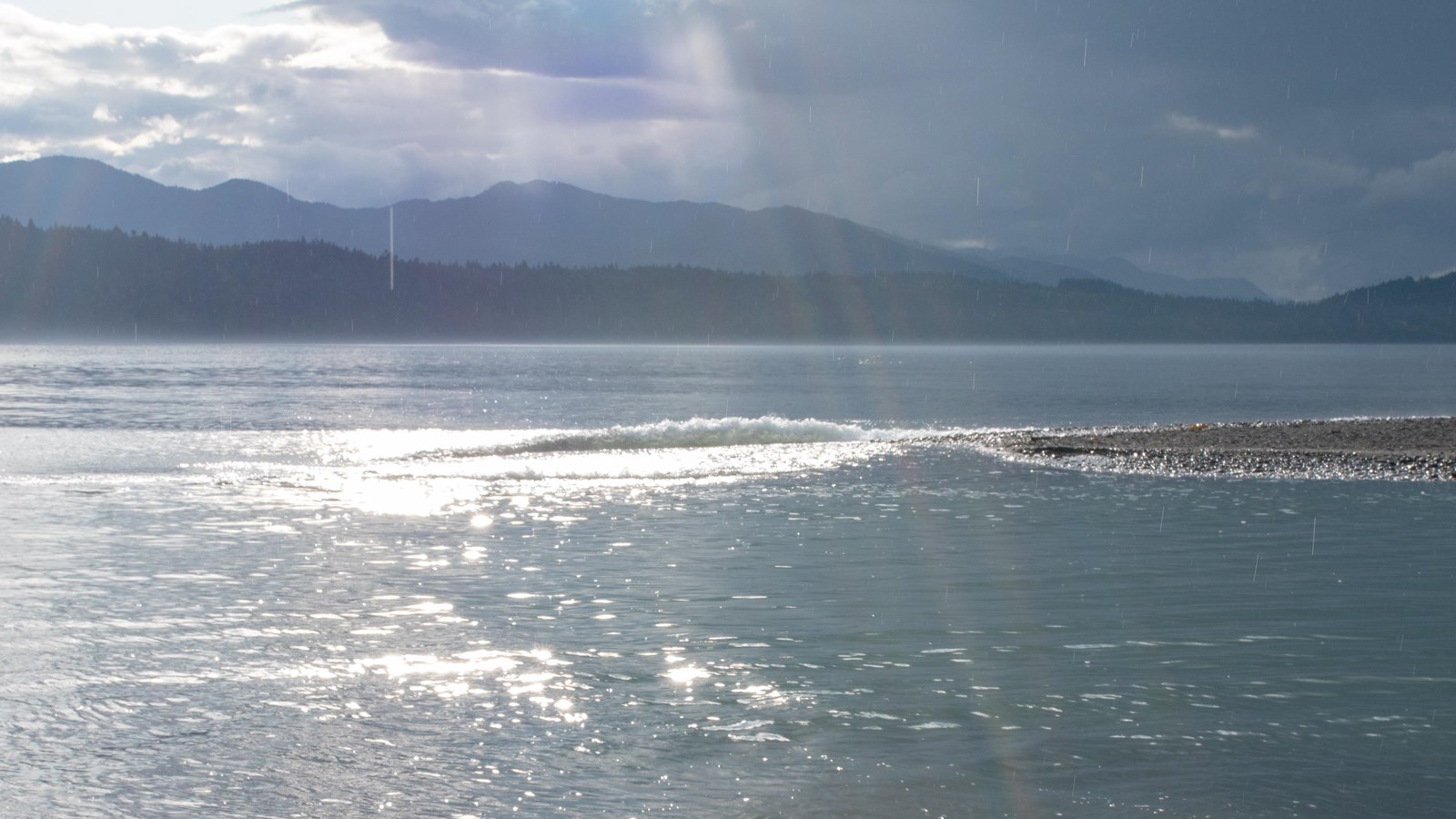 A shimmering body of water, with a gravel bar visible off shore and mountains past the far shore.