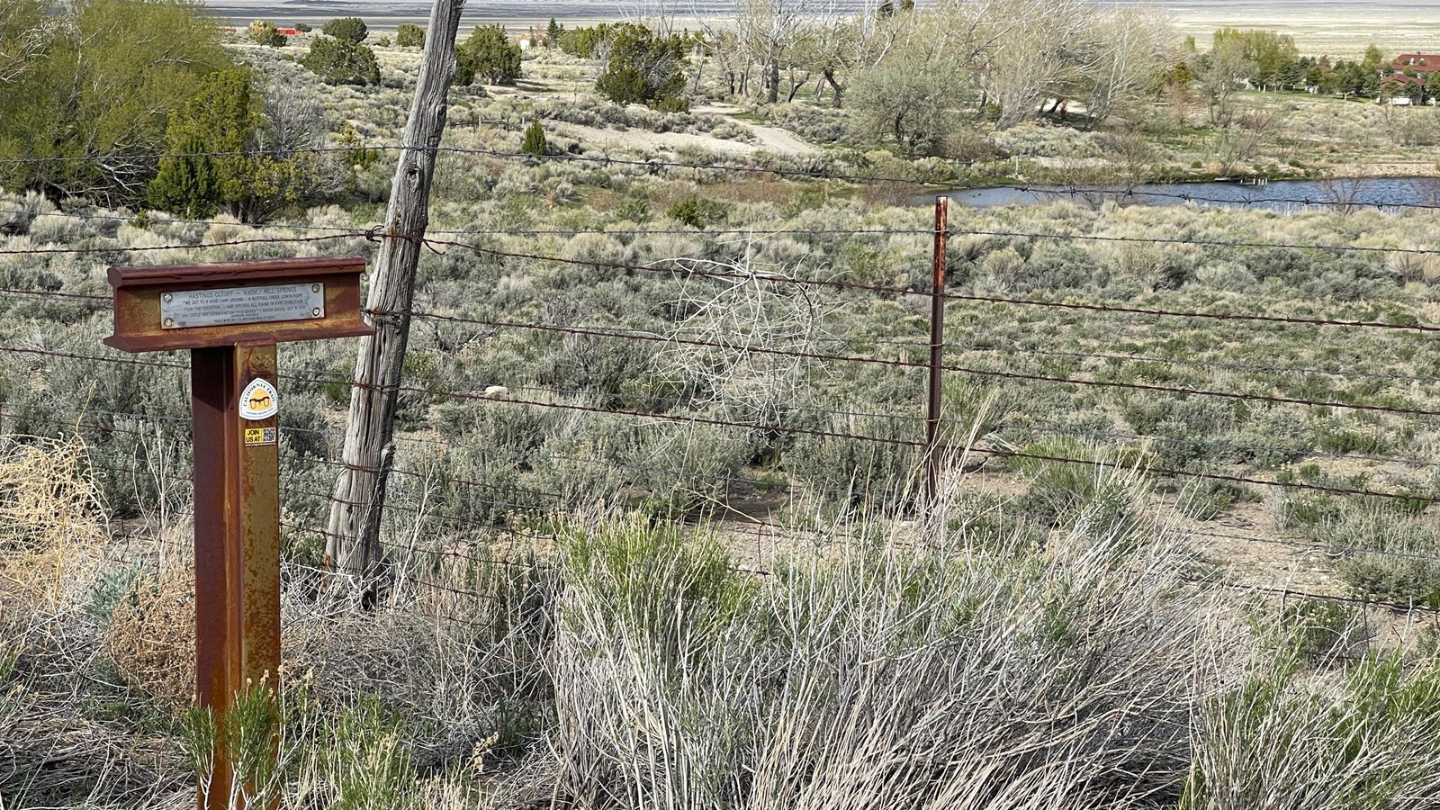 A brown trail marker stands at a barbed wire fence, surrounded by half-dead rabbitbrush. 