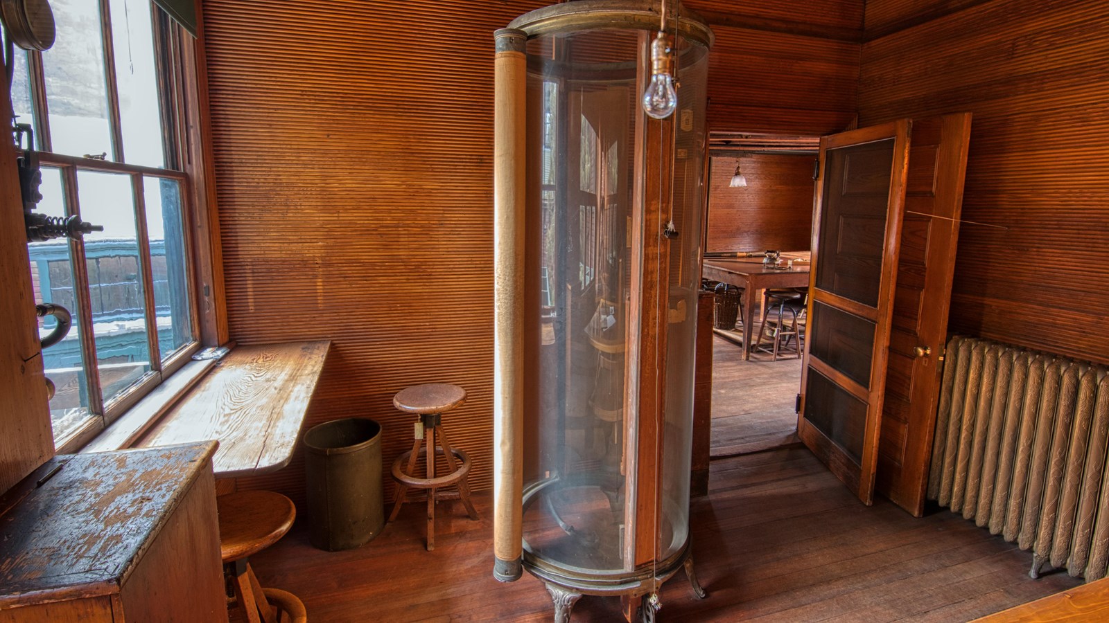 Wooden room with glass tube standing in the middle. There is a desk by the window.