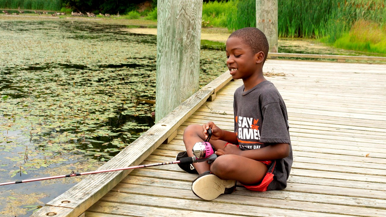 A boy sits on the edge of a dock fishing in a pond. 