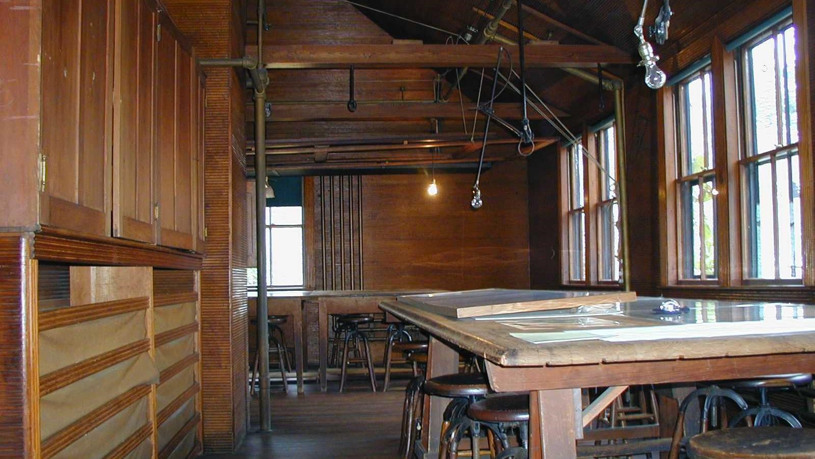 Wooden room with large table with papers on top. Lights hang down from above.
