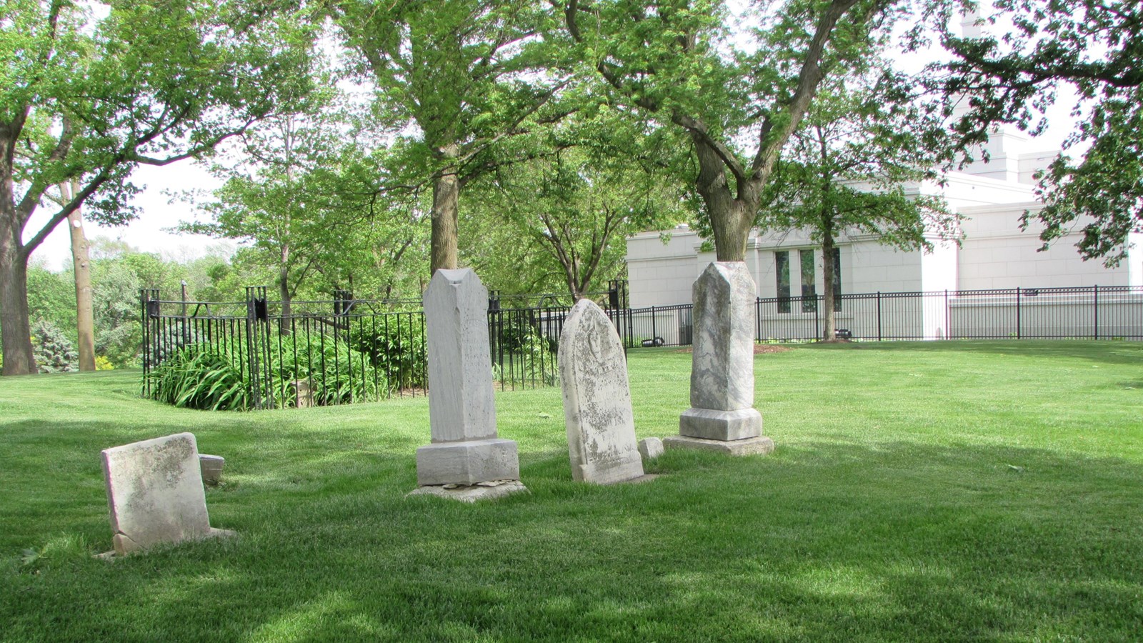 Row of four headstones in treed cemetery, well maintained lawn. Mormon Center building in background