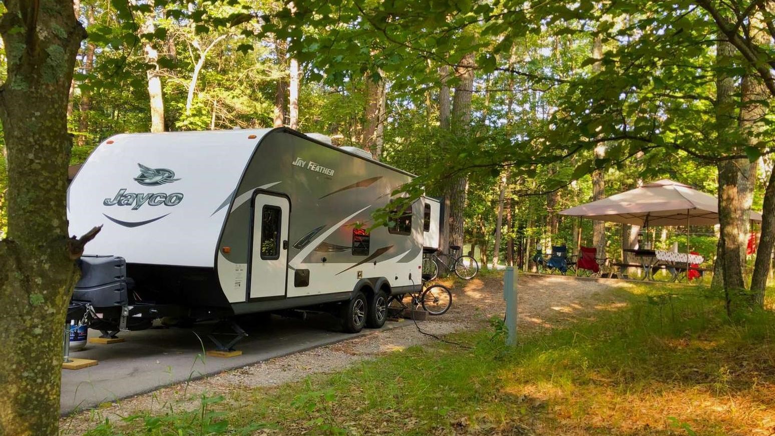 White and gray travel trailer sits on a concrete pad in a wooded campsite