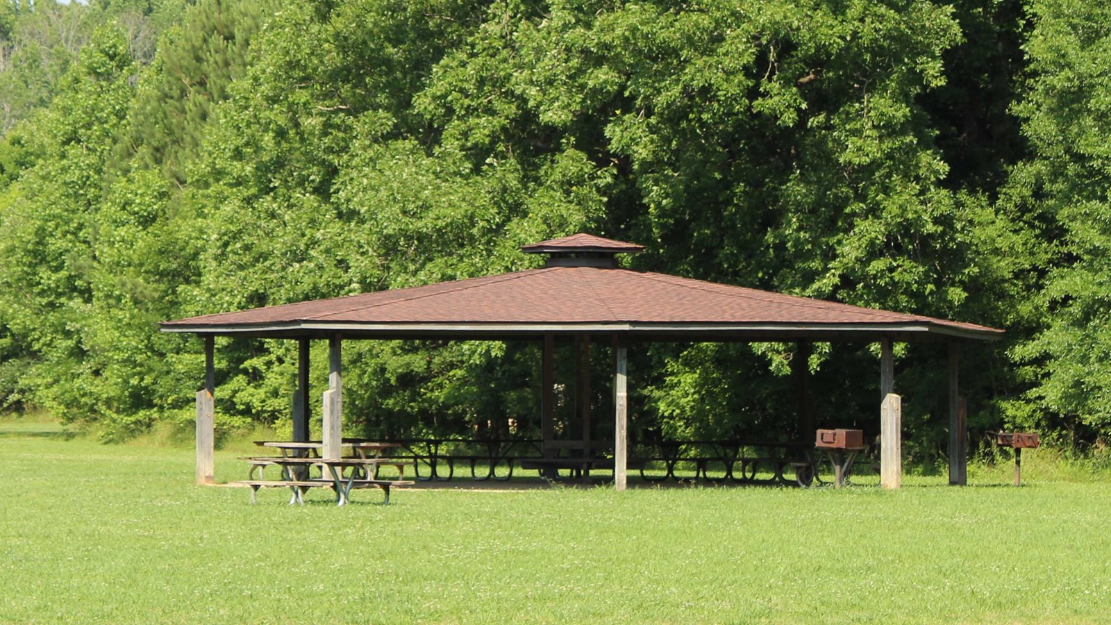 Pavilion with picnic tables and grill with field in foreground and woods in background.