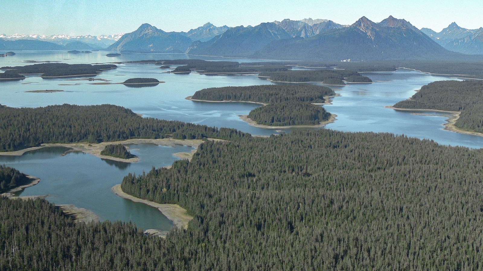 Aerial view of the beardslee islands. Many forested flat islands lie before distant tall mountains.