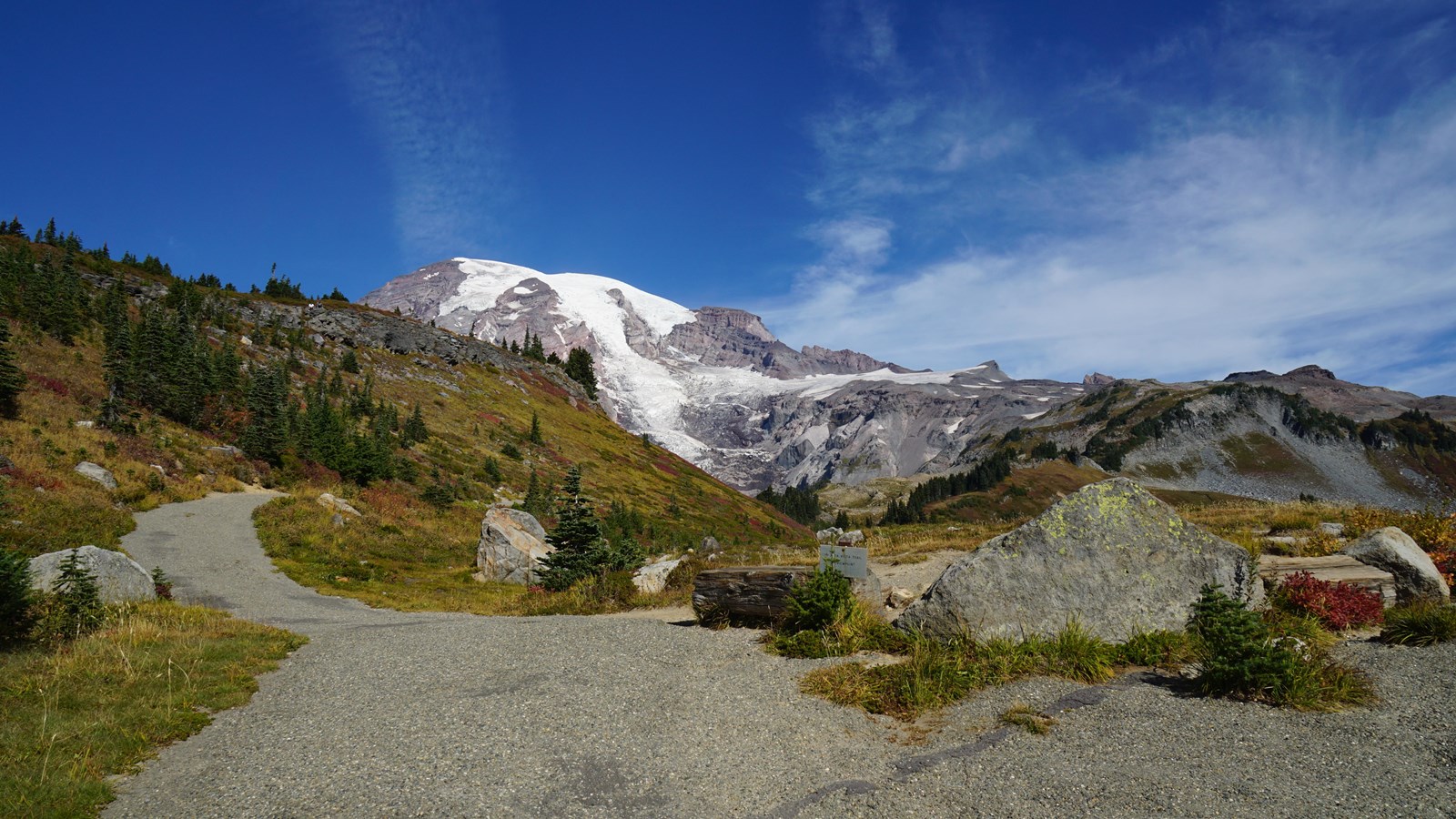 A pointed boulder is set between two paved trails. One trail climbs a ridge towards a mountain peak.