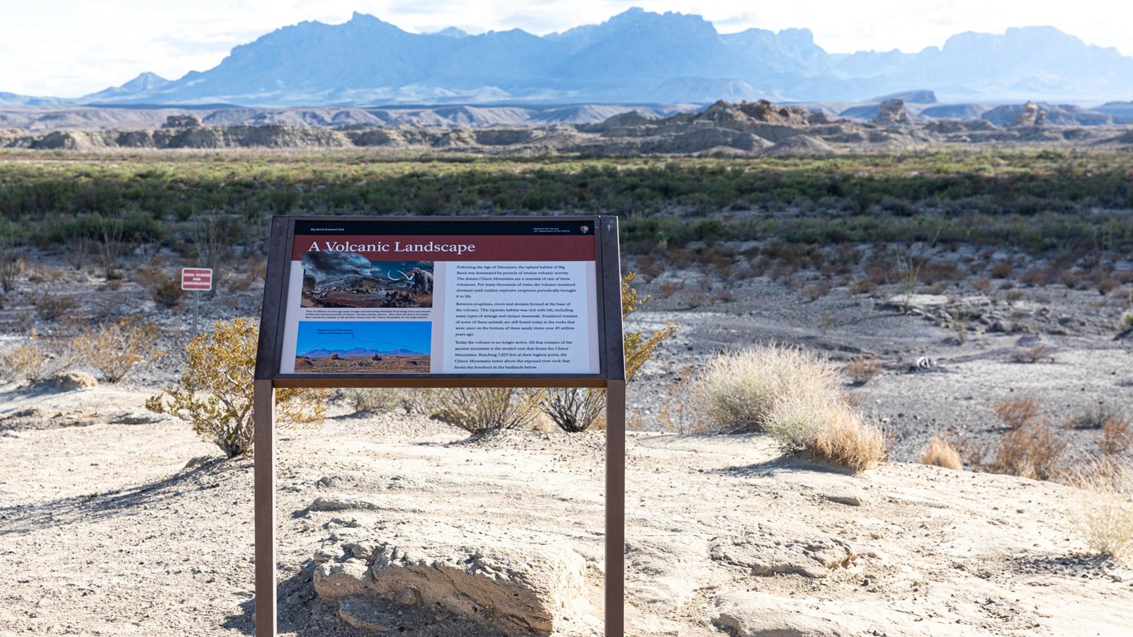 A sign supported by a metal frame is situated at the top of a small hill, overlooking a desert scene