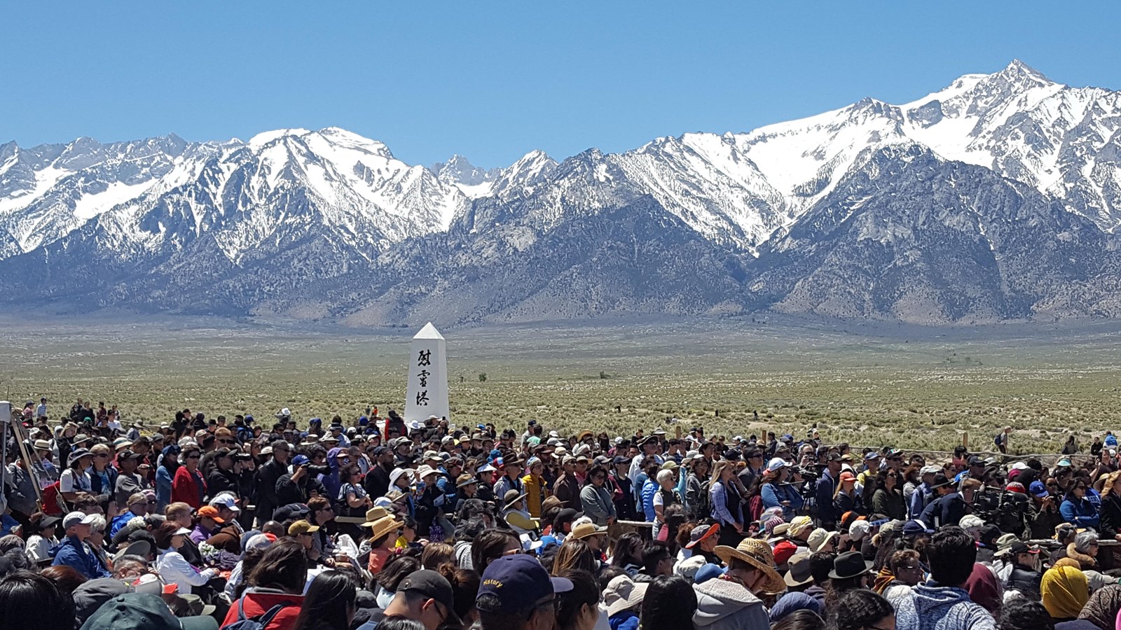 Colorful photo of a large crowd surrounding the Manzanar Cemetery Monument in the sun.