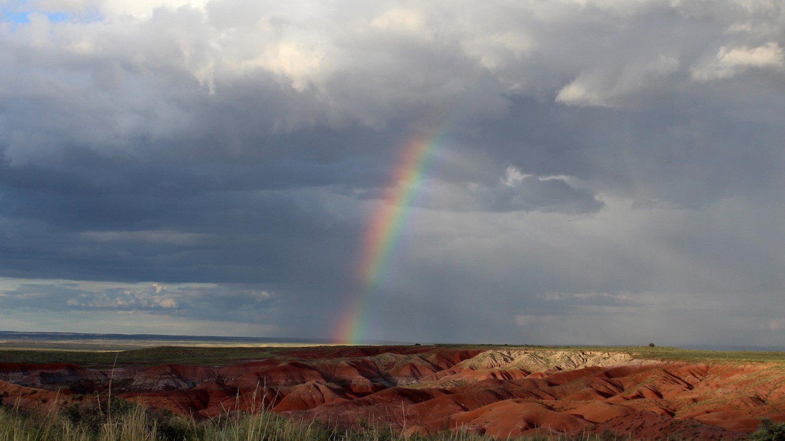 Red badlands and grassland with cloudy sky and rainbow.