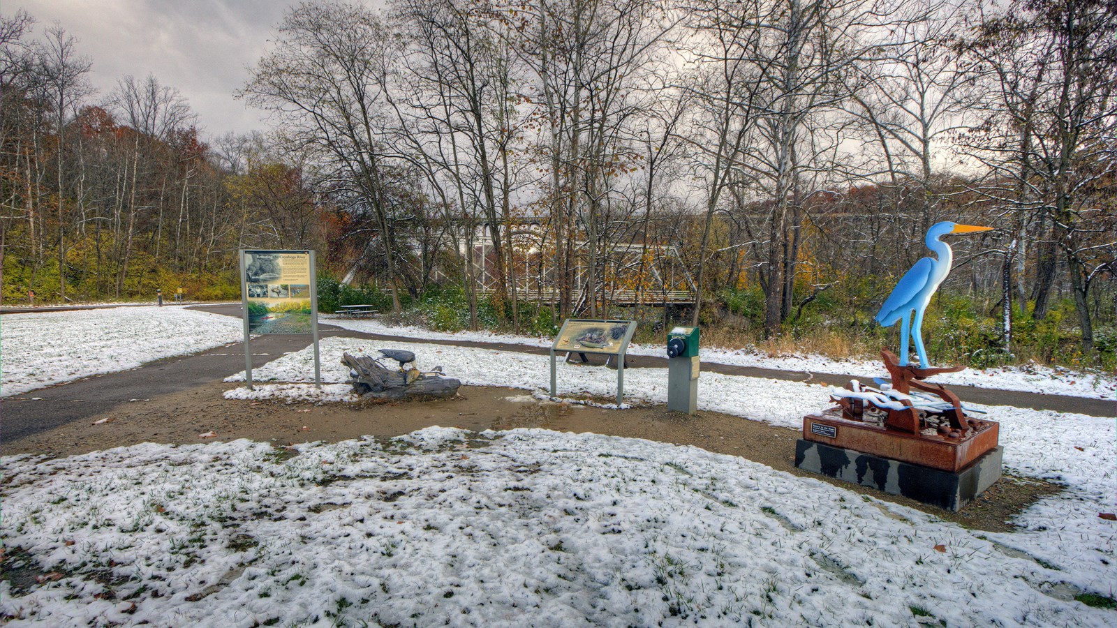 Two graphic panels, statue of eagle on a log, & tall blue heron statue beside paved trail in snow.