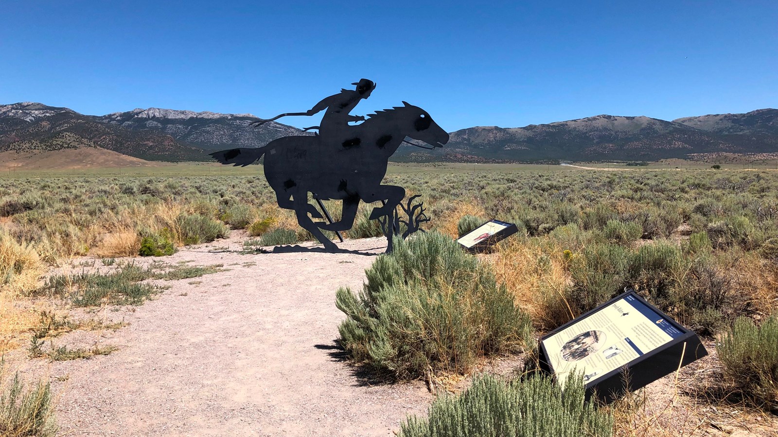 A gravel path with wayside exhibits leads to a metal silhouette cutout of a Pony Express rider.