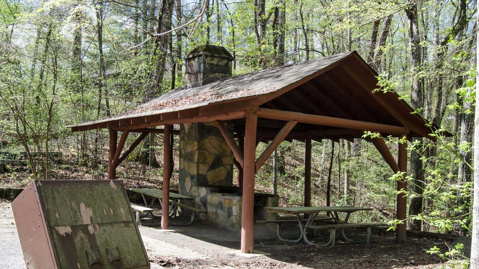 Rustic shelter with a massive stone grill and two picnic tables in a wooded area.