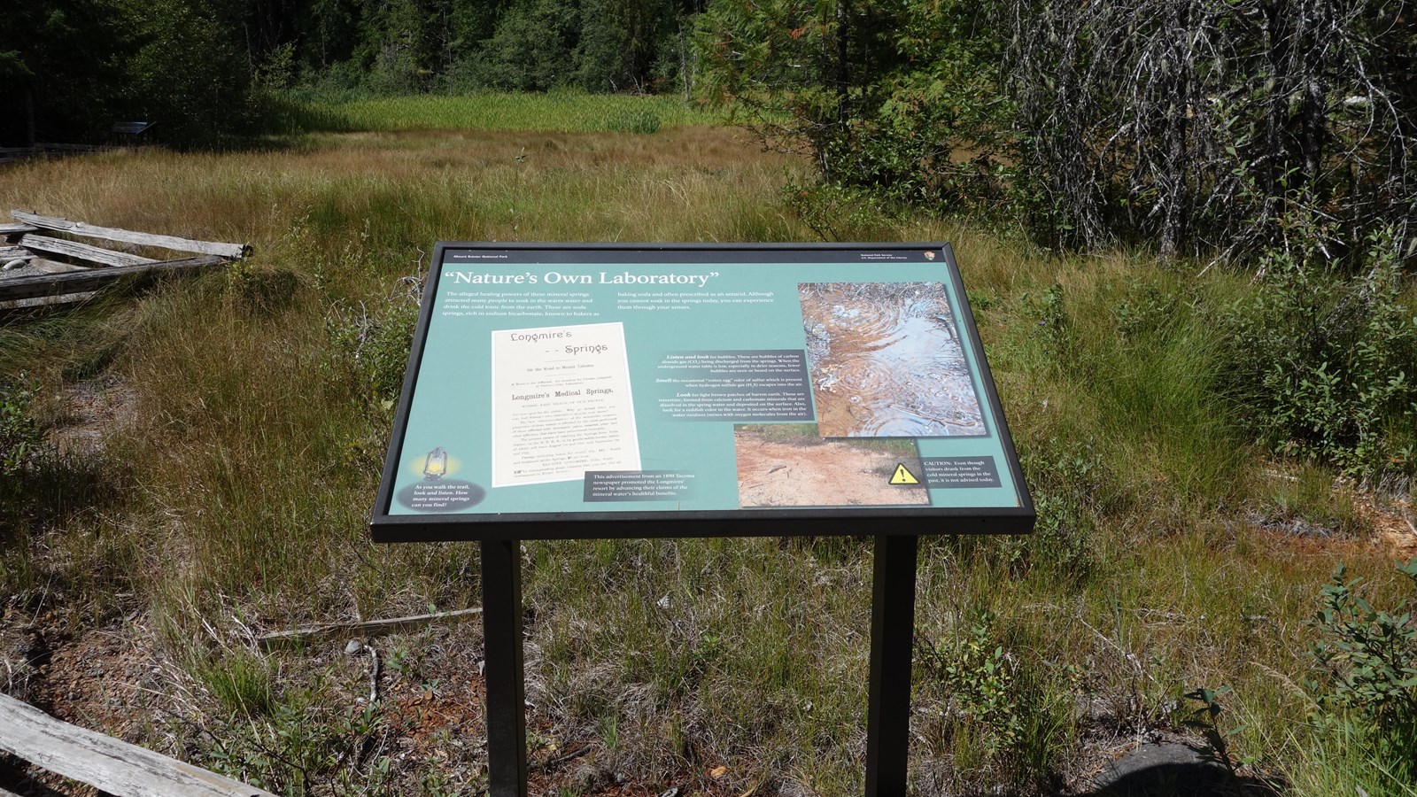 A wayside in a metal stand next to a trail and split-rail fence in a section of meadow.