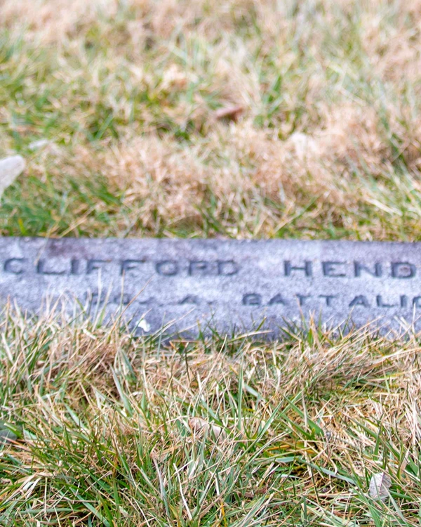 A dark colored gravestone engraved with the name Clifford Henderson lays flat among grasses.