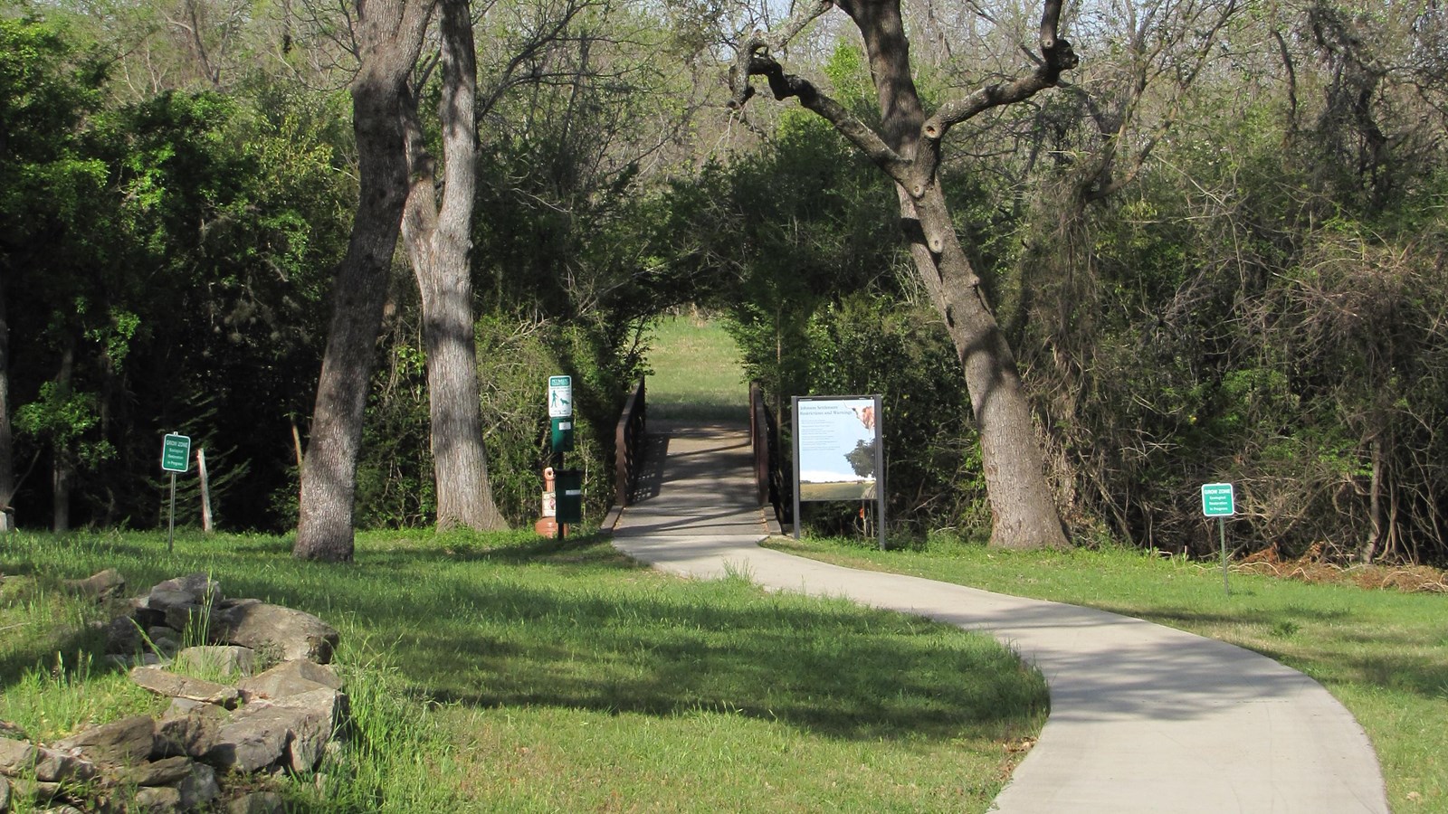 A concrete path bends towards a break in the green foliage under tall trees.