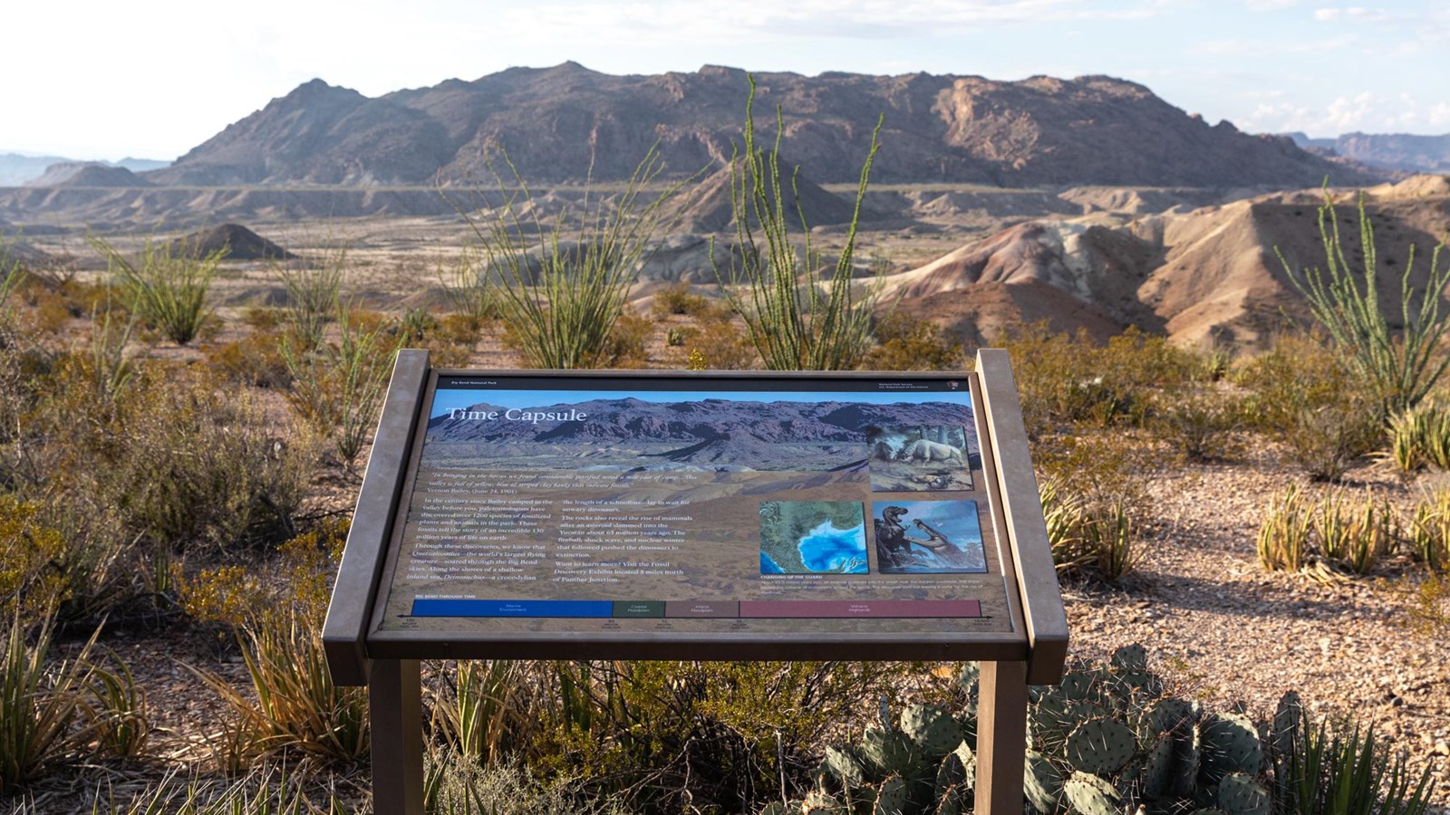 A metal sign atop a hill overlooks colorful clay hills situated in a wide drainage.