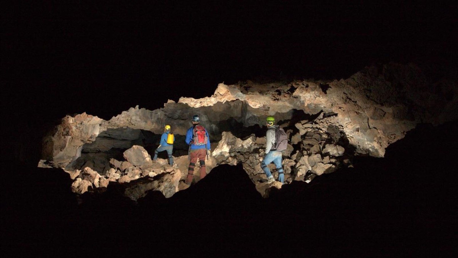 Cavers walk over boulders and rubble in a dark lava tube cave