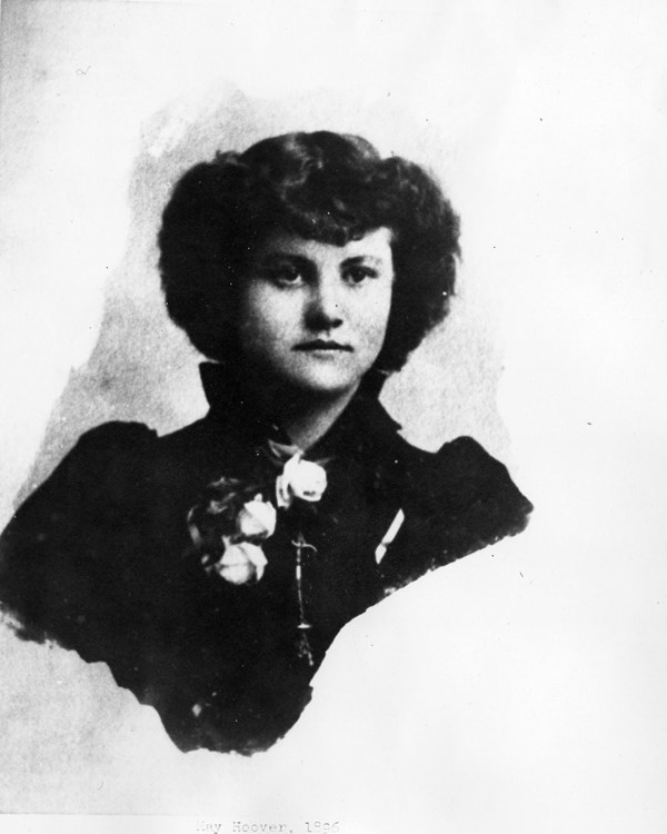 A portrait photo from 1896 depicts a well dressed young woman with flowers pinned to her dress.