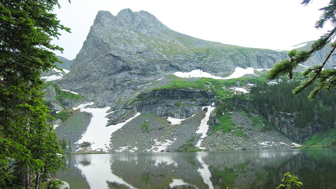 An alpine lake with conifer trees at the side, and rocky summit with snow 