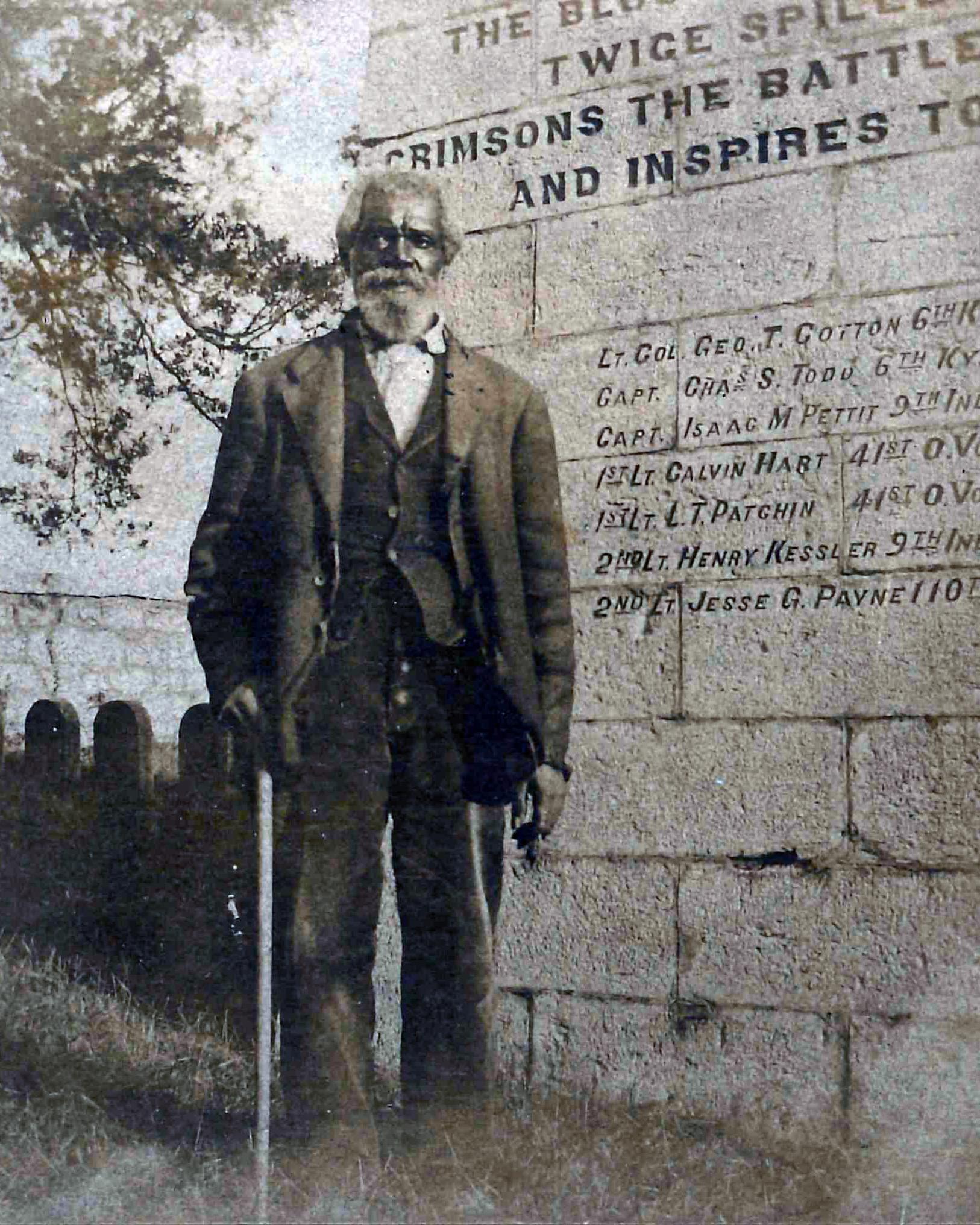 An elderly Black man in a suit stands next to a cube shaped monument.