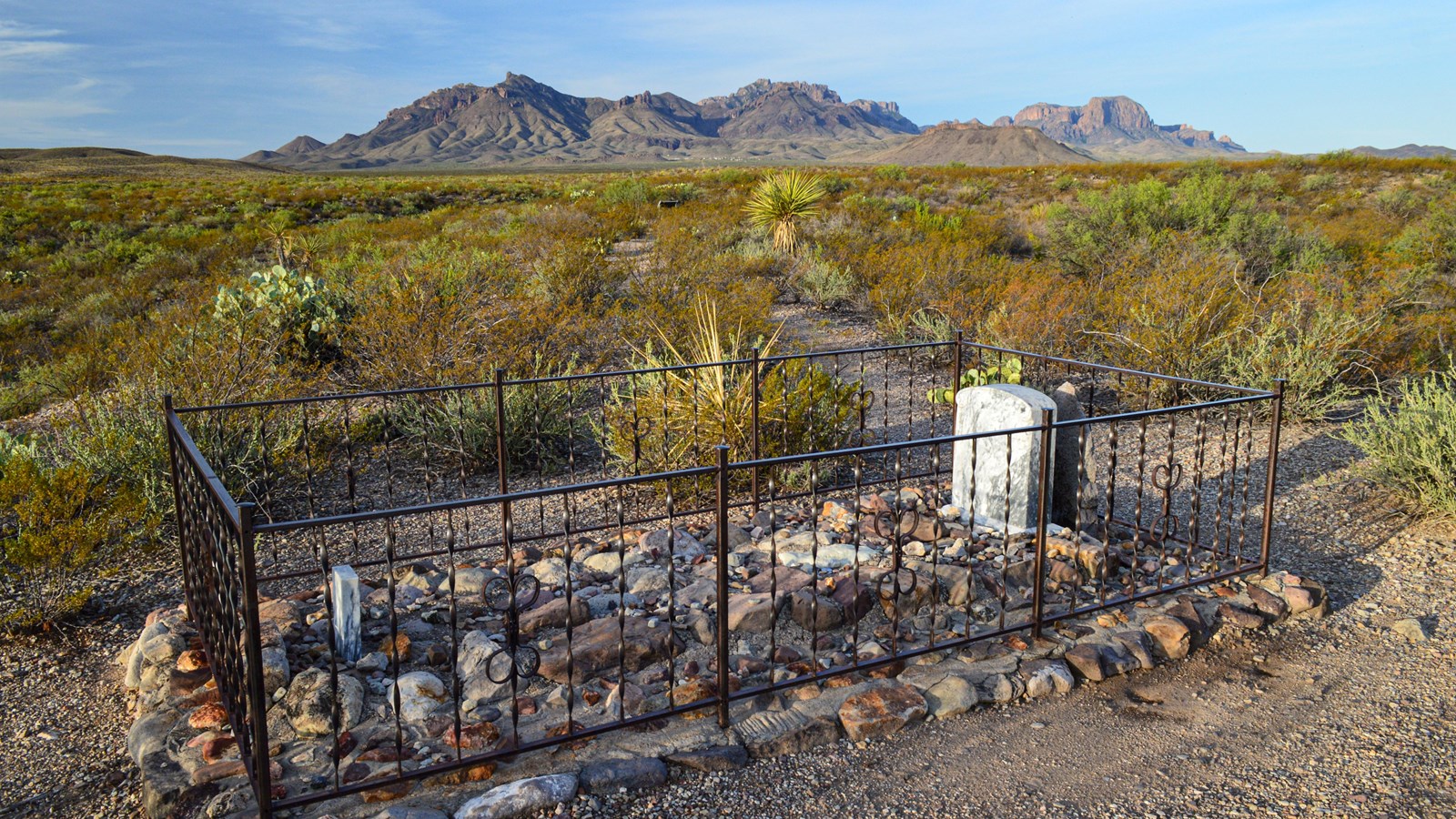 A metal fence surrounds a gravesite covered with large rocks with a granite headstone.