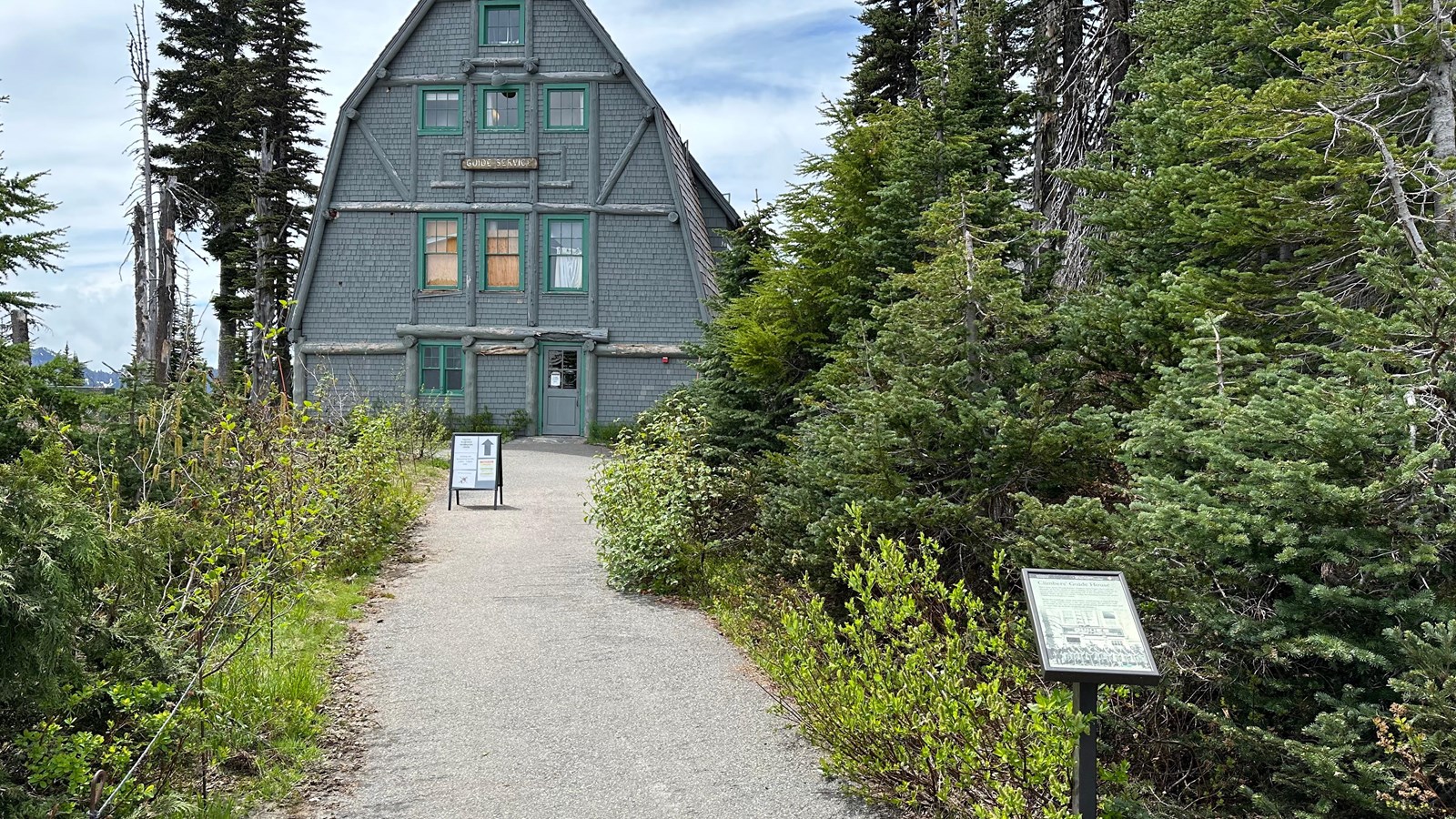 A small exhibit panel next to a paved path leading to a tall wood building with a steep angled roof.