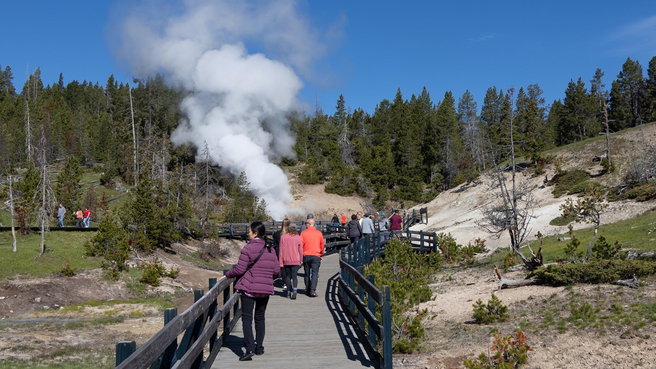 People walk down a boardwalk towards a large plume of steam.