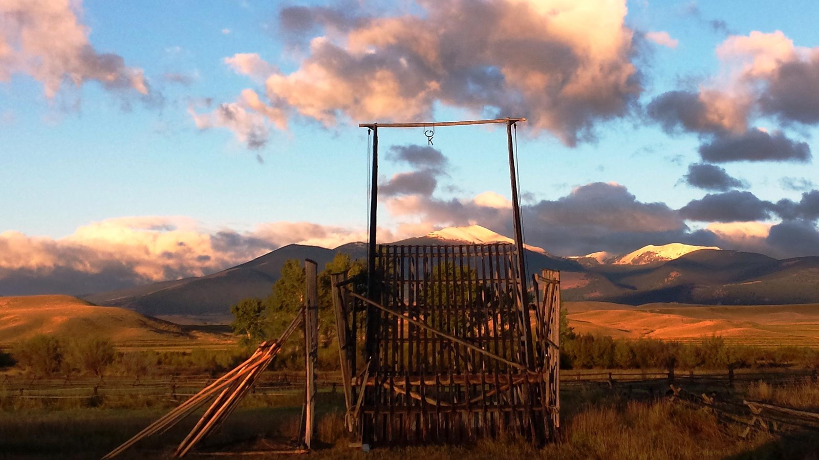 A beaverslide hay stacker sits in a grassy field. In the distance is a row of trees and mountains.