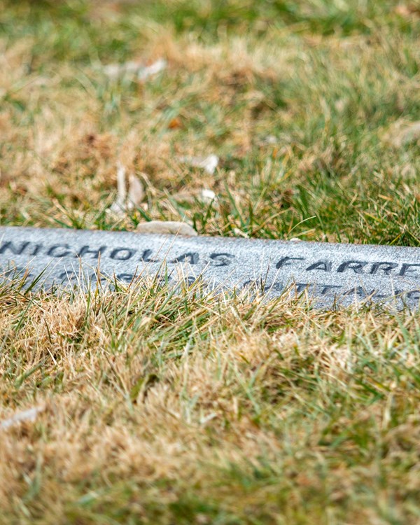 A dark gravestone is carved with the name of Nicholas Farrell lays flat on the ground among grass.