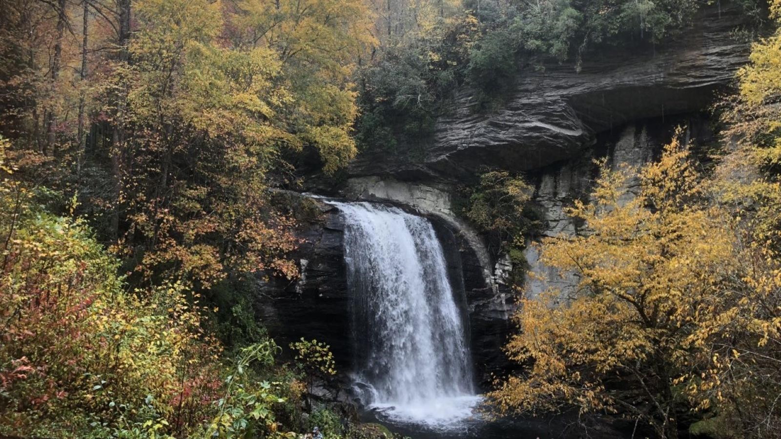 Tall waterfall cascades over rock shelf to a pool of water below with fall colors in background.