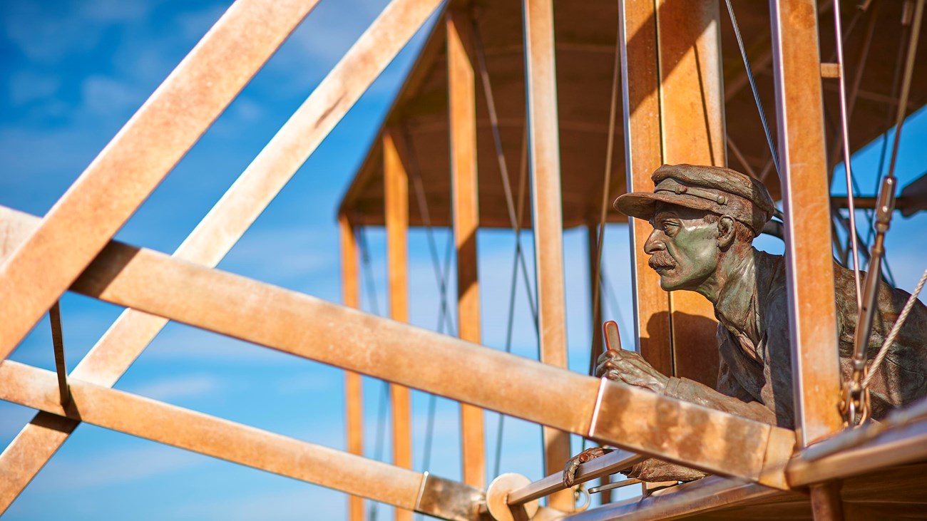 A bronze sculpture of Orville Wright surrounded by wooden beams of the flyer