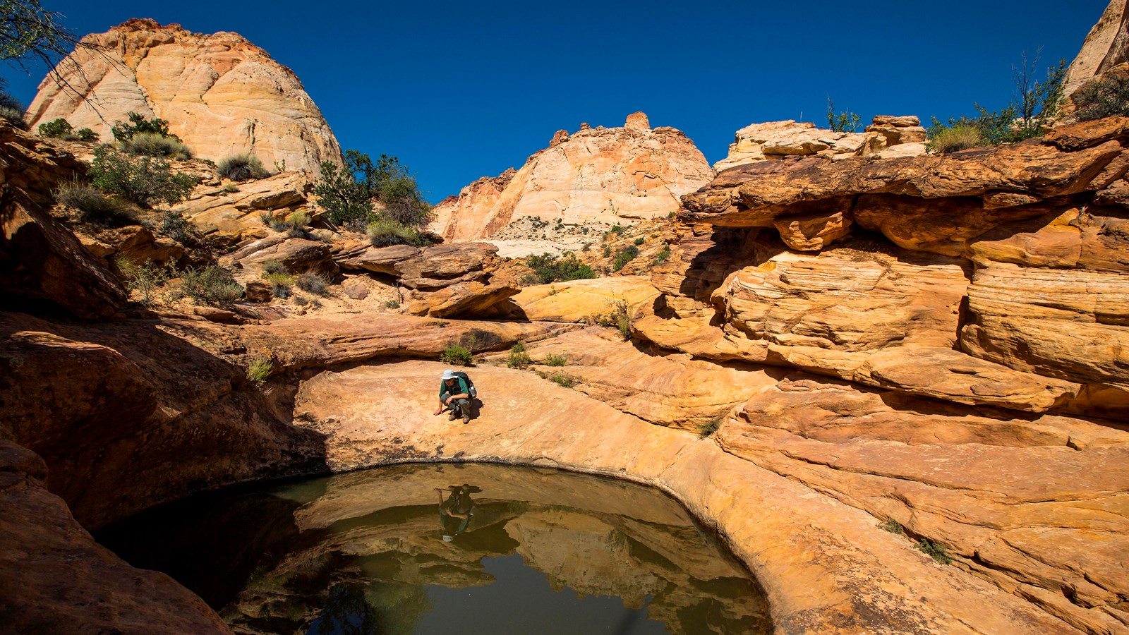 A man crouches near a still pool of water in tan colored sandstone.