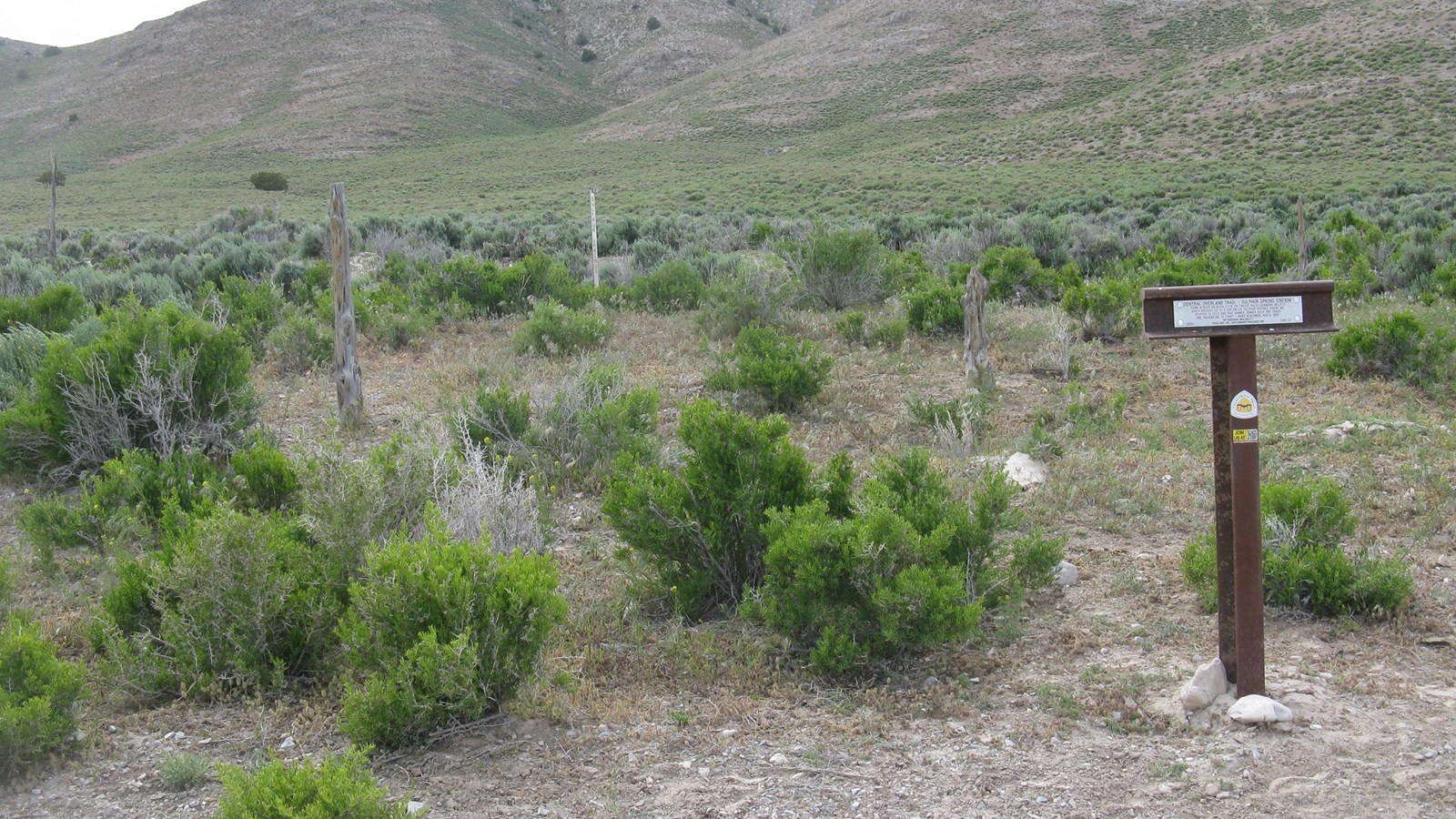 A brown, T-shaped post stands outside a barbed wire fence, with hills rising in the background. 