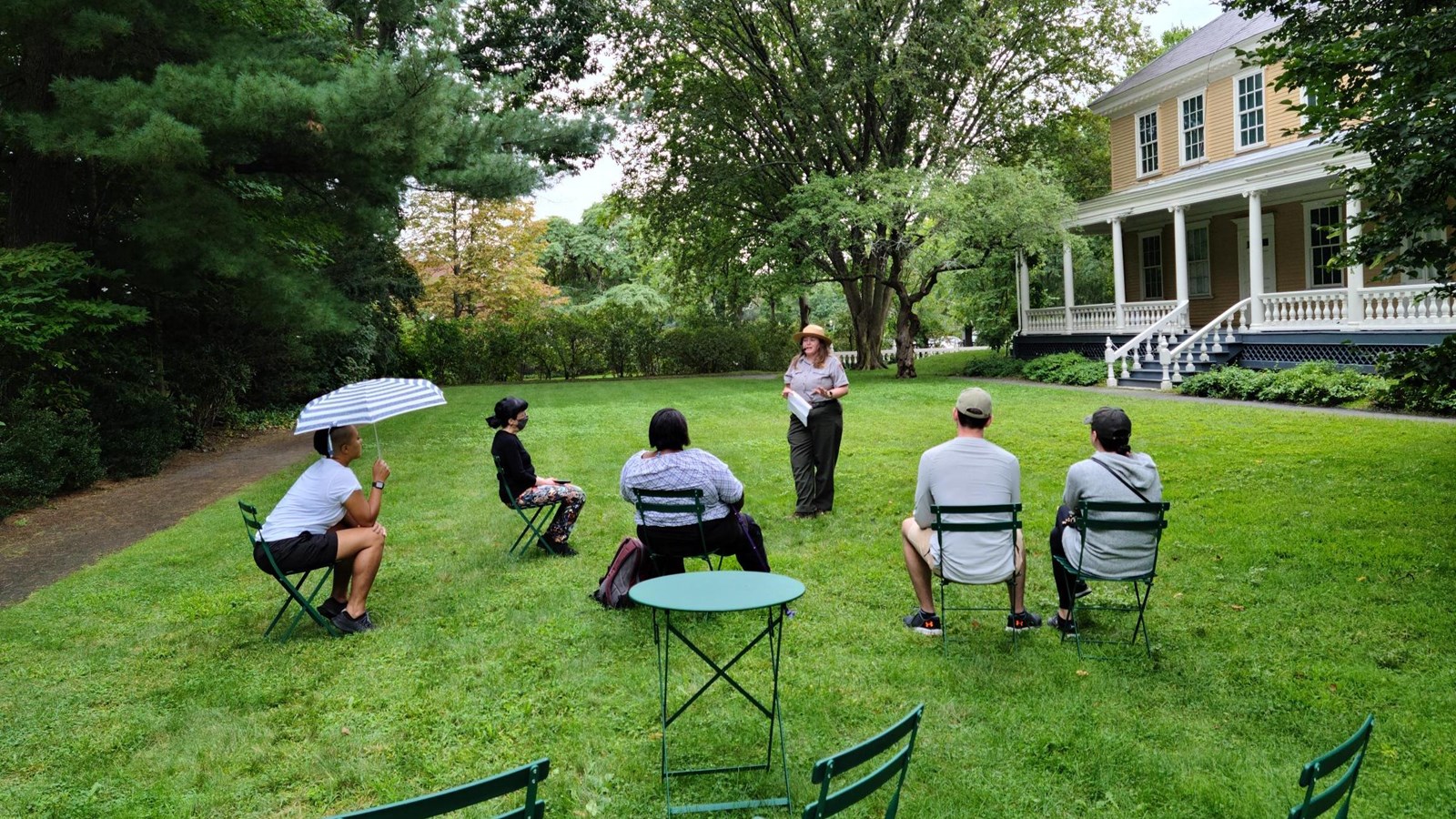Standing ranger talking with five seated visitors on a green lawn with trees and house in background