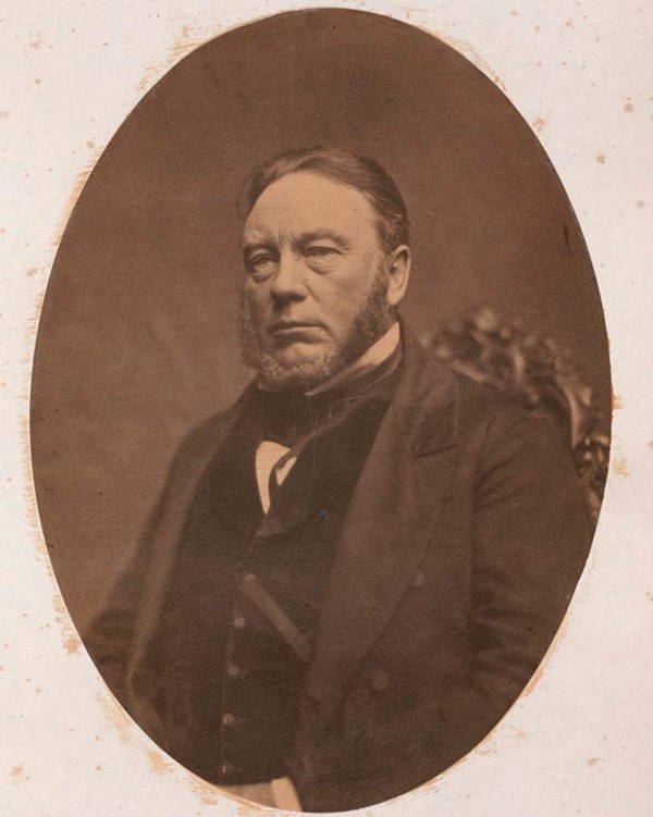white middle-aged man wearing a threepiece suit. He has a thick chinstrap beard & a blank expression
