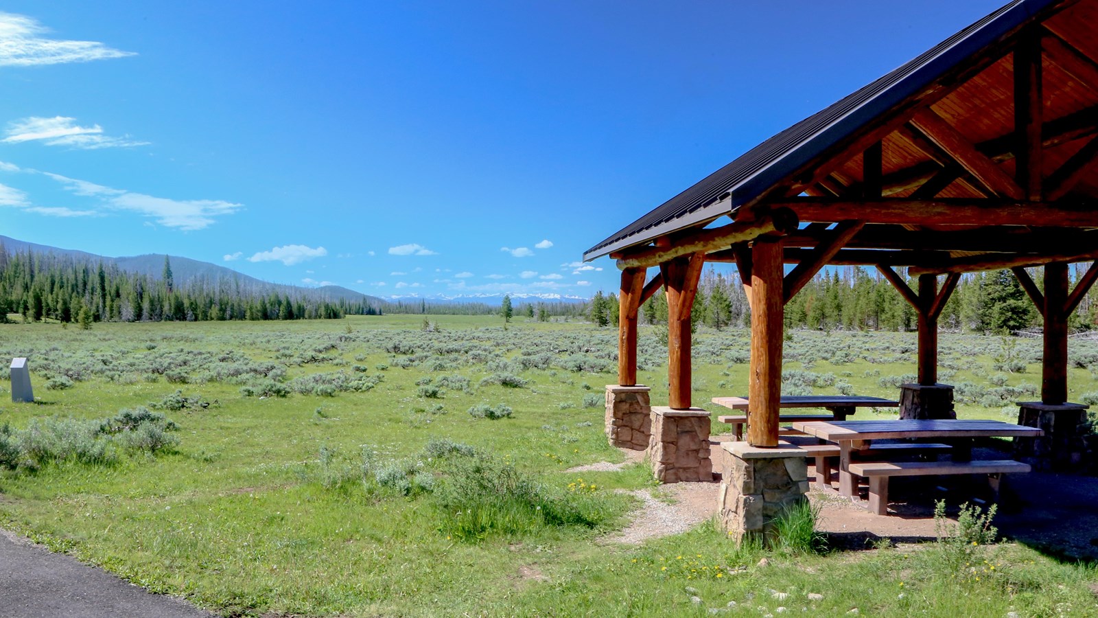 A wooden picnic shelter opens onto a broad, green field under a blue sky.