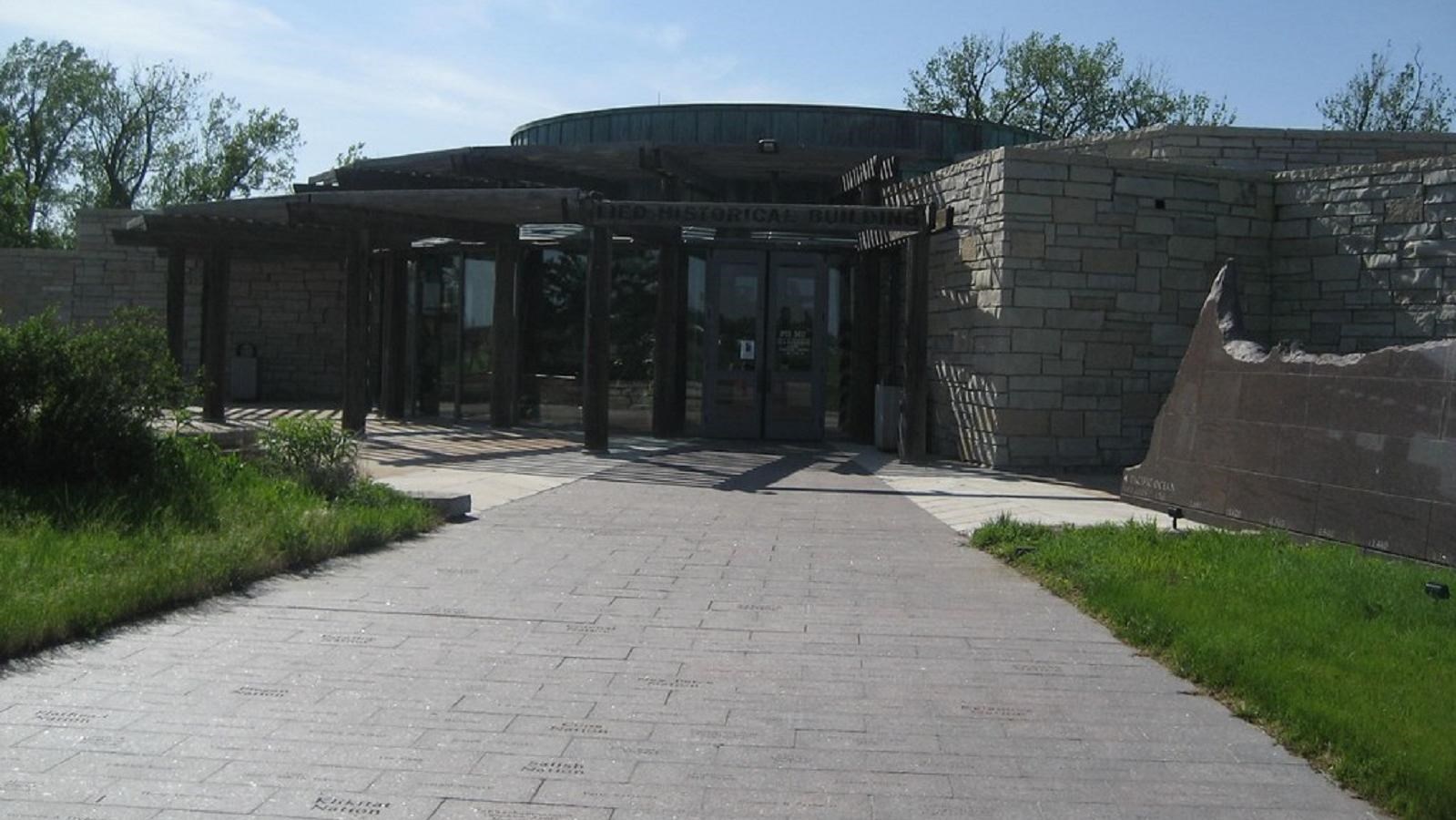 A low-lying building of gray stone with glass doors flanked by trees sits on a green lawn.
