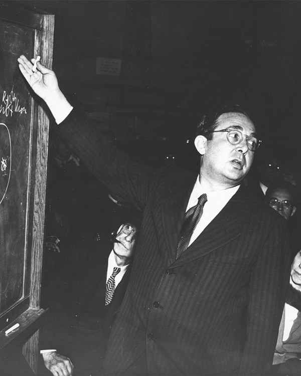 Black and white photo of a man gesturing to a chalkboard.