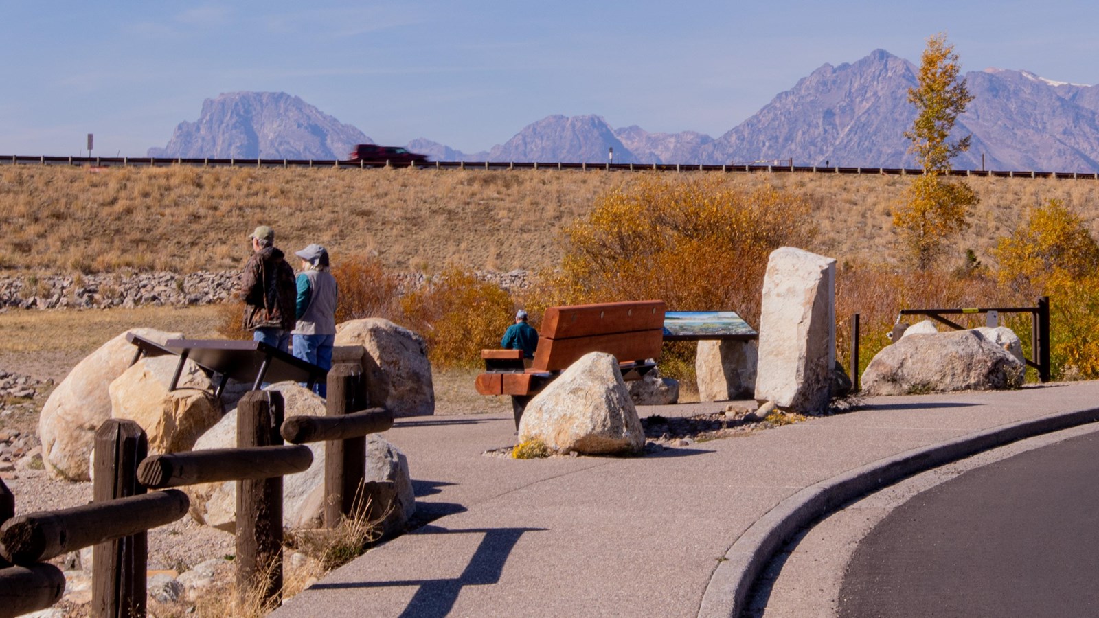 Two visitors look towards the dam (out of picture) standing by interpretive panels and a bench.